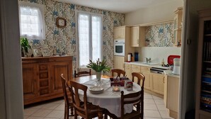 A dining area with a wooden table set for four, surrounded by chairs. There is a vase with flowers on the table, and a clock on the wall. - Cottage for 7 people 15 minutes from Puy du Fou (Pouzauges)