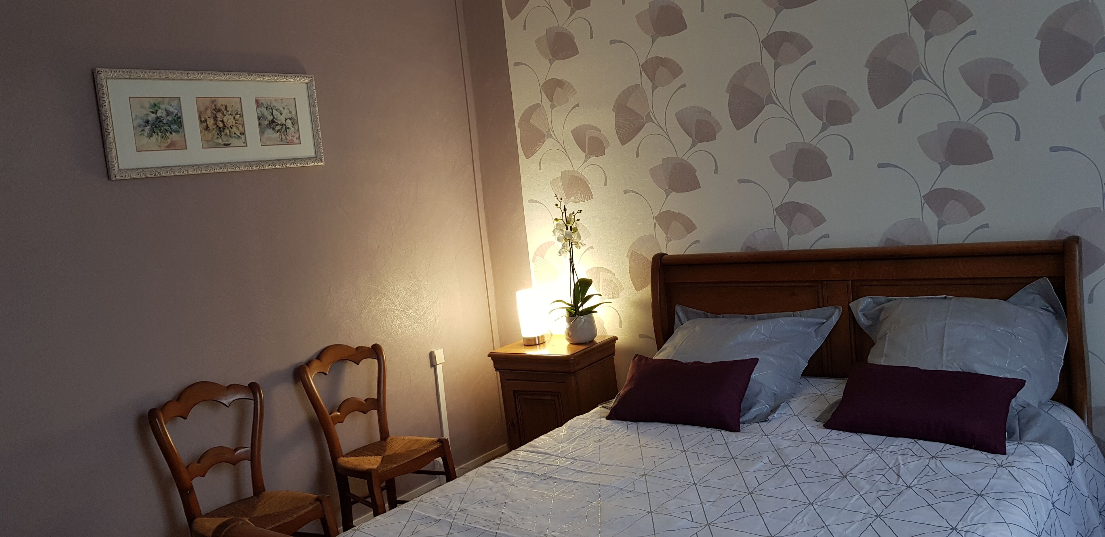 A bedroom with a wooden headboard, a bed with patterned bedding, a nightstand with a lamp and a potted plant, and a chair.