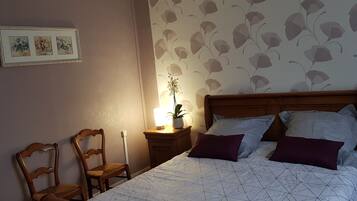 A bedroom with a wooden headboard, a bed with patterned bedding, a nightstand with a lamp and a potted plant, and a chair.