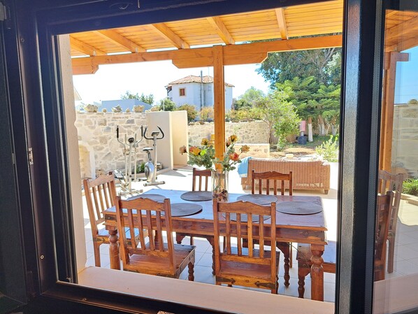 A wooden table and chairs under a pergola, with outdoor exercise equipment and a stone wall in the background.