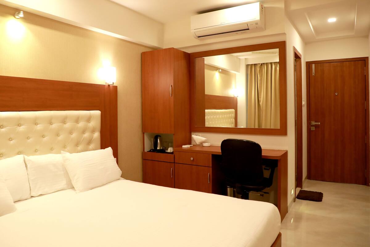 A hotel room with a large bed, a wooden desk, a chair, a mirror, and an air conditioning unit.