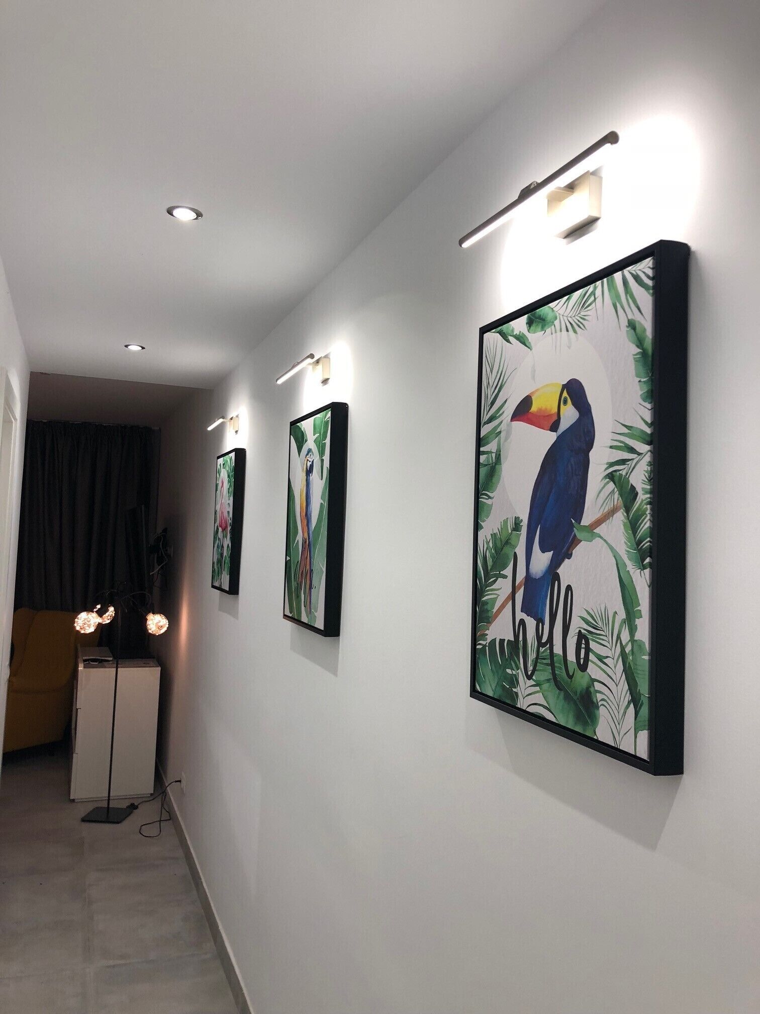 Een gang met drie kunstwerken in lijsten aan de muur, een wit plafond met inbouwverlichting en een kleine tafel met een lamp.