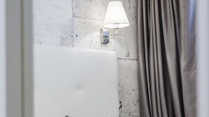 Un letto rifatto con cura, con una testiera bianca, cuscini grigi e color acquamarina, e una coperta grigia. Una tenda grigia è parzialmente socchiusa.