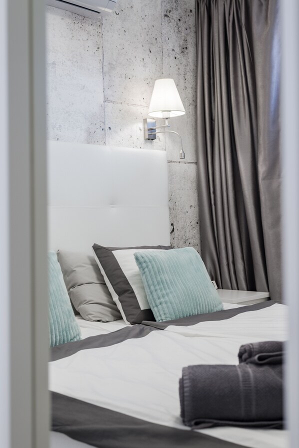 A neatly made bed with a white headboard, grey and teal pillows, and a grey blanket. A grey curtain is partially drawn to the side.