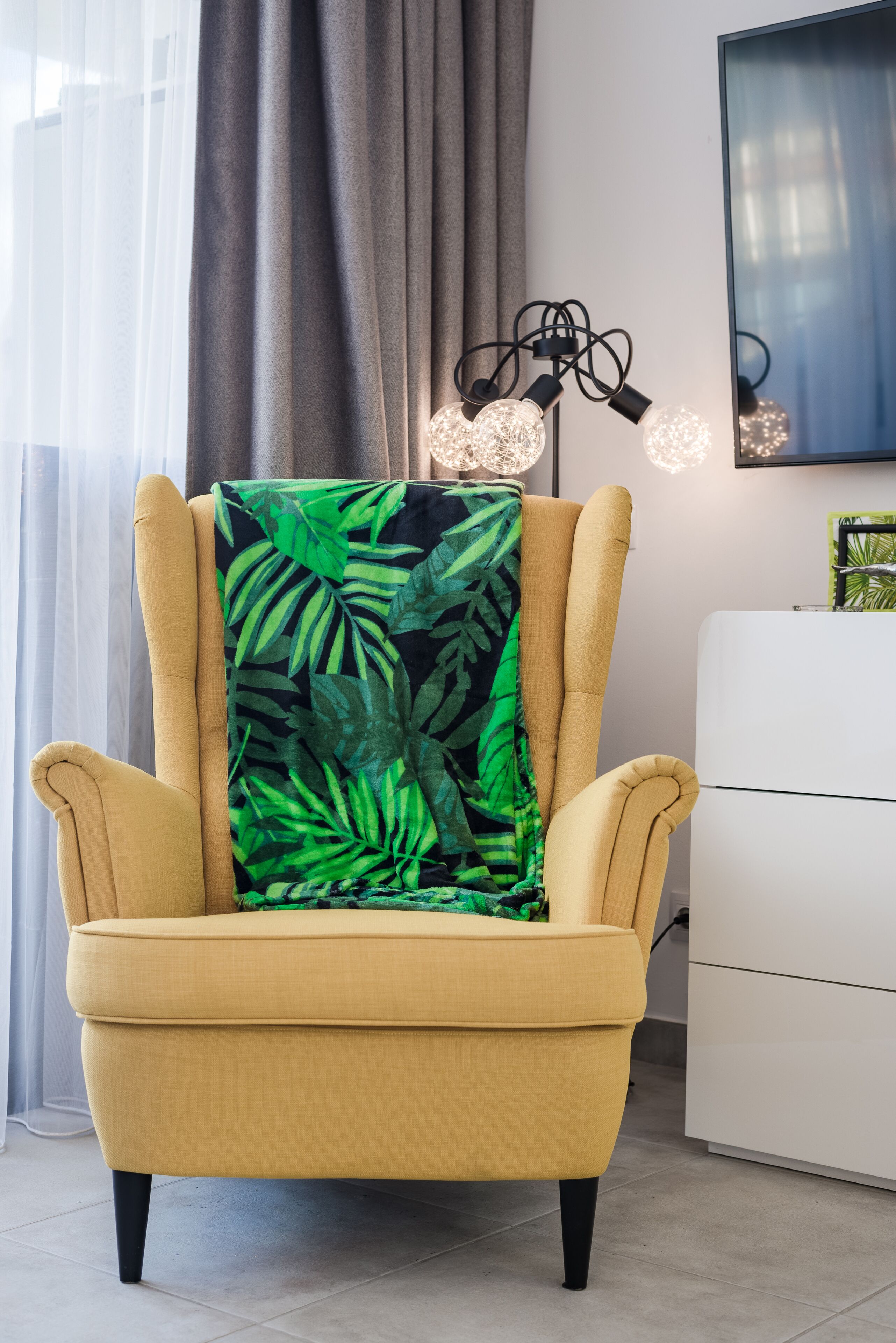 Un fauteuil jaune agrémenté d’un plaid orné de motifs de feuilles tropicales, un luminaire moderne et un meuble blanc dans une pièce au style contemporain.