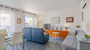 A modern living room with a sofa, a dining table with chairs, a TV, and a small kitchen area. - Royal Marine by Interhome (Fréjus)