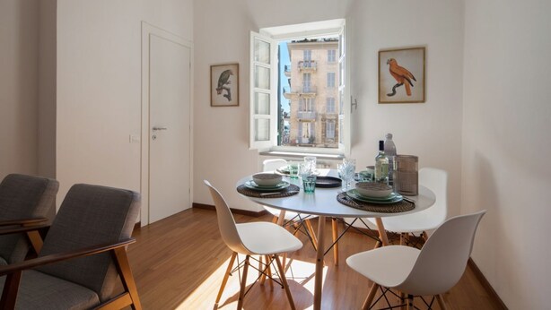 Een eetgedeelte met een tafel gedekt voor vier personen, twee stoelen aan weerszijden, en een raam met uitzicht op gebouwen.