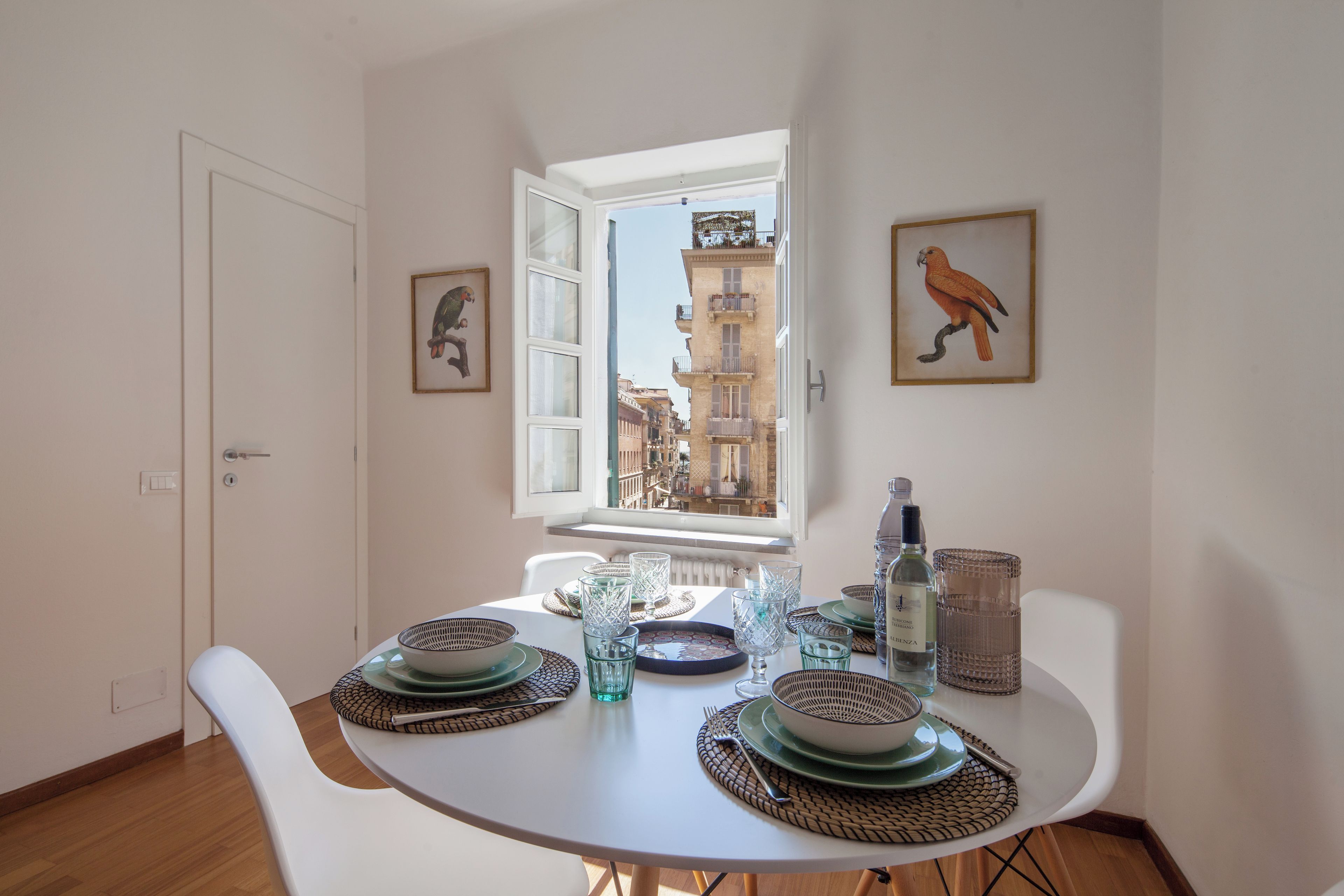 Un'area pranzo con un tavolo apparecchiato per due, una finestra che si affaccia su edifici e due quadri appesi al muro.