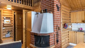 A rustic cabin interior with a brick fireplace, wooden walls, a wooden dining table, and a kitchen area. - Jänkäluppo 1 by Interhome (Äkäslompolo)