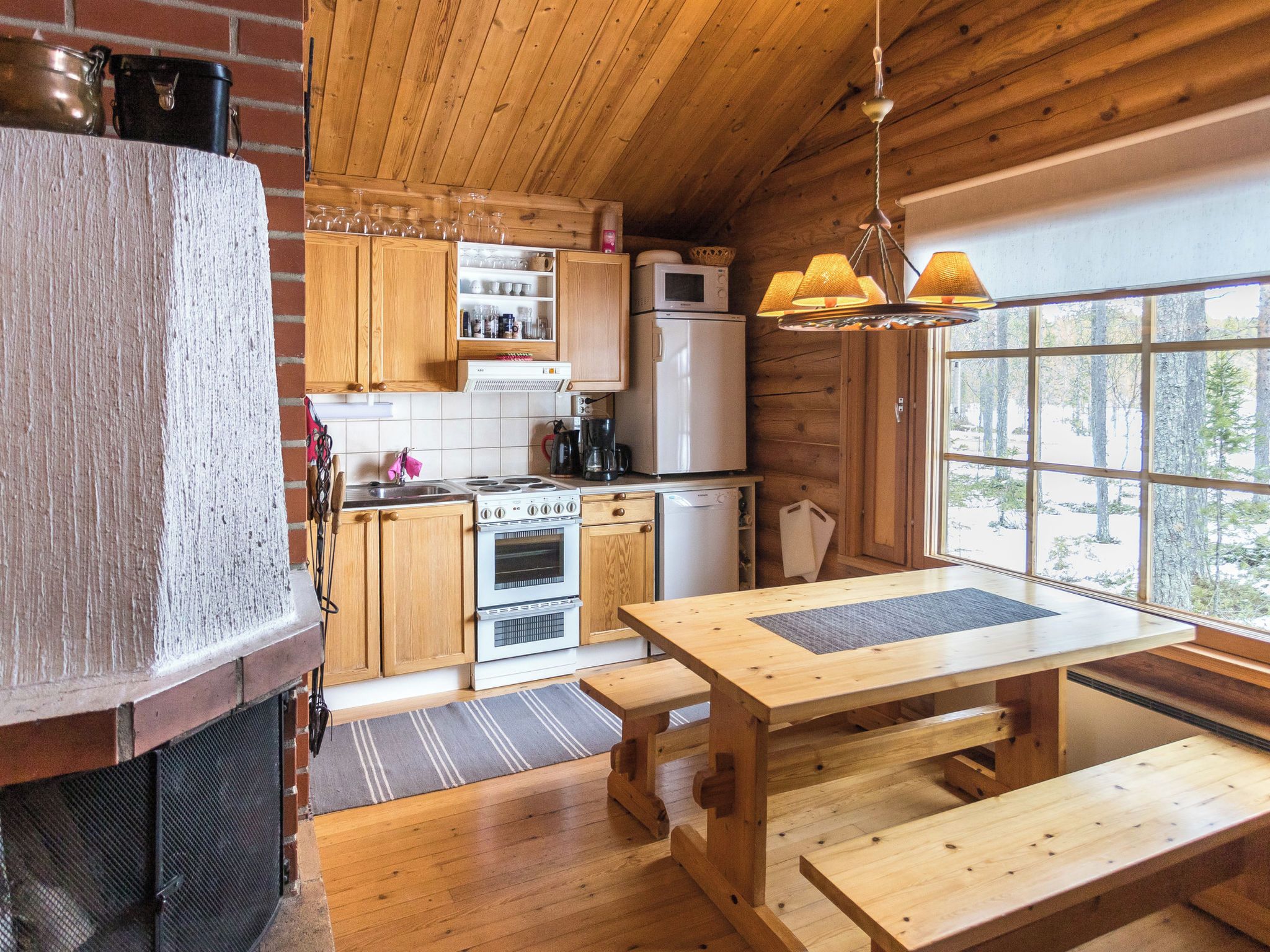 Une cuisine rustique dans un chalet, avec des meubles en bois, un poêle, un four à micro-ondes et une grande fenêtre donnant sur une forêt.