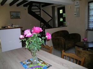 Un coin repas avec une table dressée pour deux personnes, un vase de roses roses et une carte posée sur le set de table.