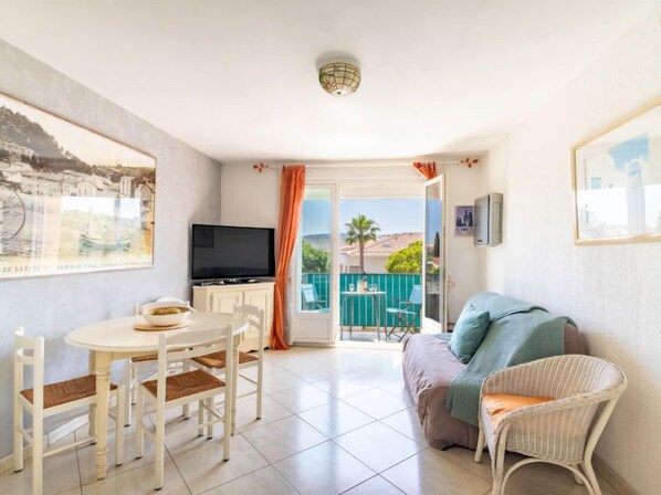 A room with a dining table, chairs, a sofa, a television, and a balcony with a view. - Les Ilaires by Interhome (Le Lavandou)
