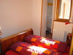 A bedroom with a wooden bed frame, a red and orange patterned bedspread, and a window with a view of a bathroom.