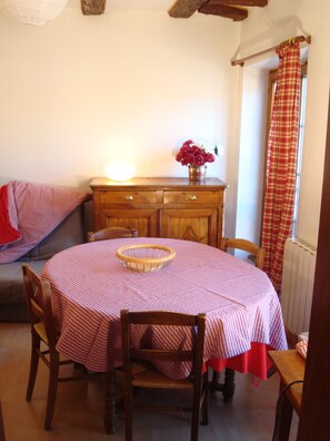 A dining area with a round table covered in a checkered tablecloth, wooden chairs, a wooden cabinet with a vase of flowers, and a window with a checkered curtain.