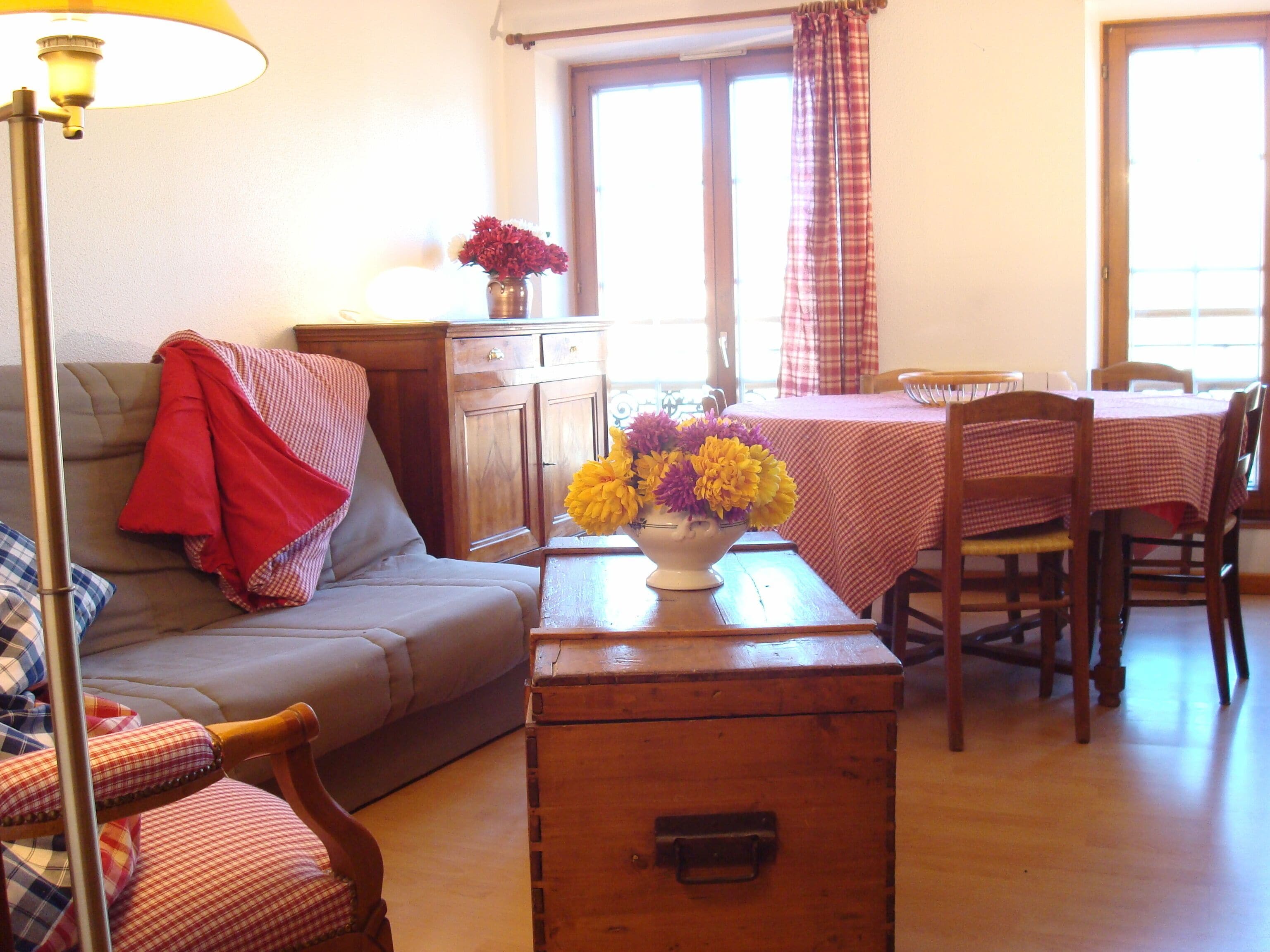 A cozy living room with a sofa, a wooden chest, a table with a vase of flowers, and a dining area with a table and chairs.