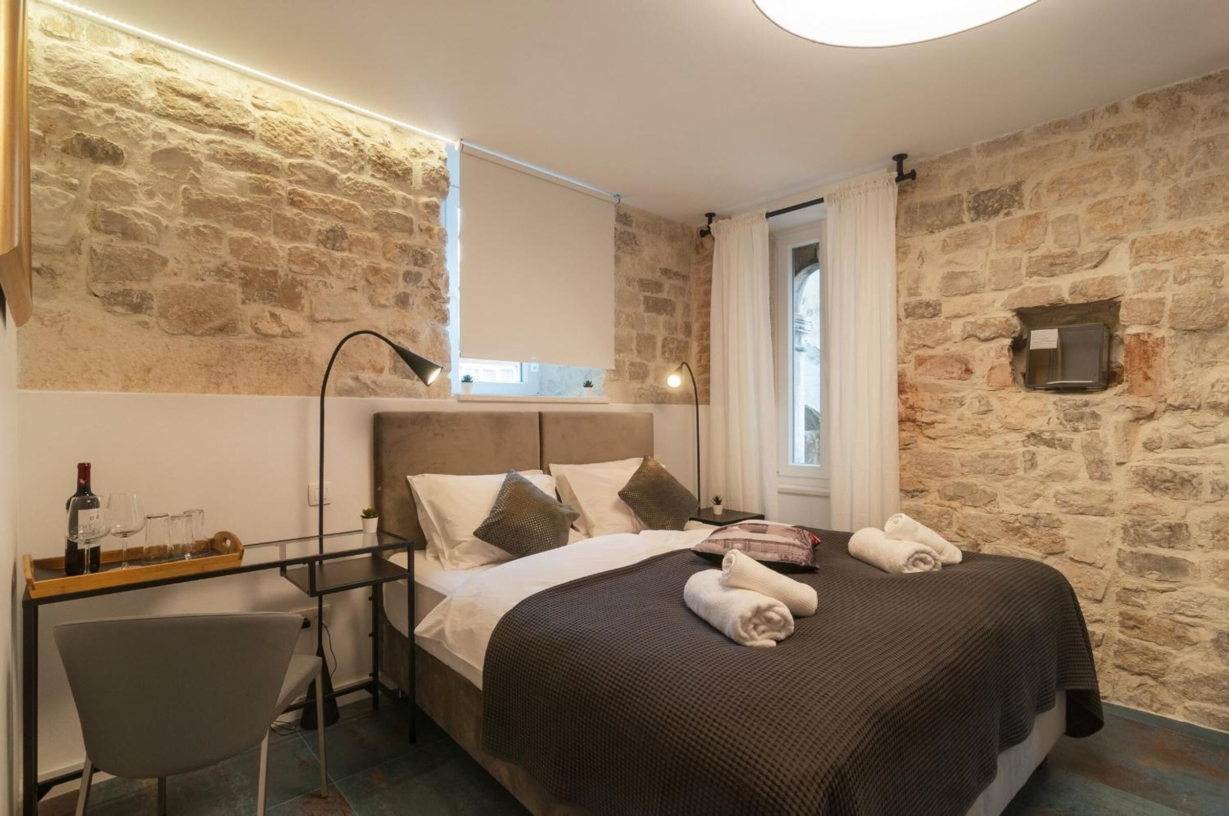 Un dormitorio con pared de piedra, una cama con manta marrón, una mesita de noche con una botella y vasos, y una silla.