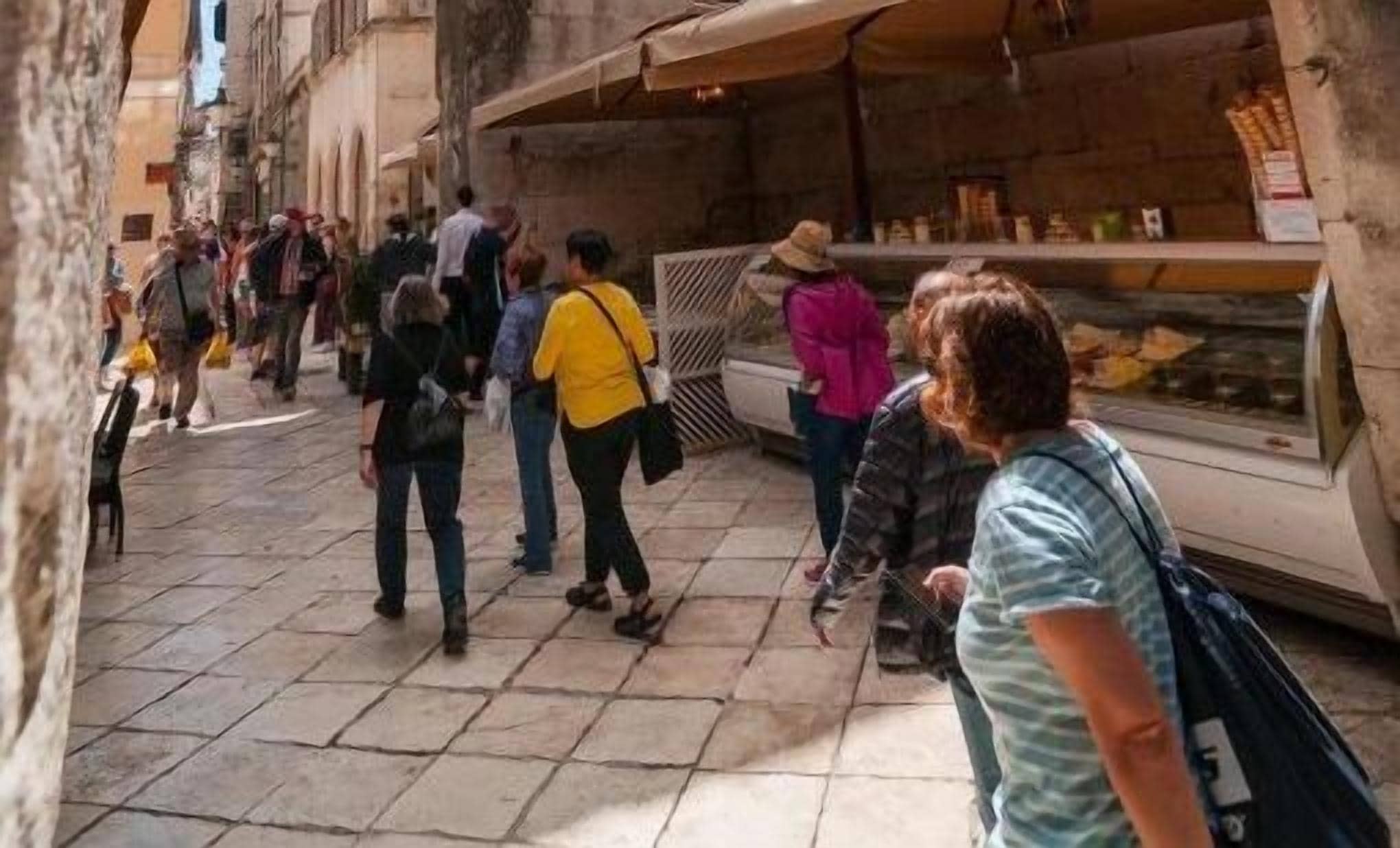 Egy keskeny utca, ahol emberek sétálnak, és a jobb oldalon egy ételárusító található.