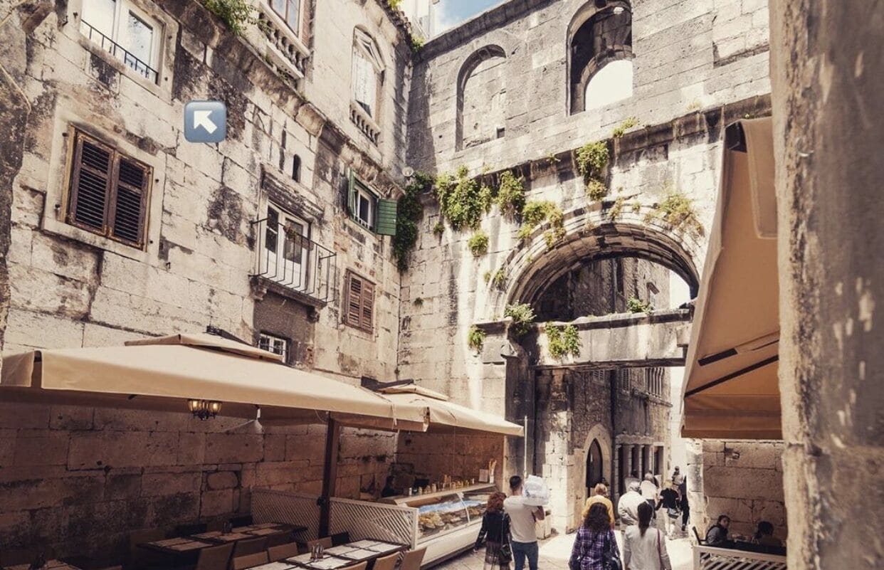 A narrow street with stone buildings, outdoor seating, and an archway.