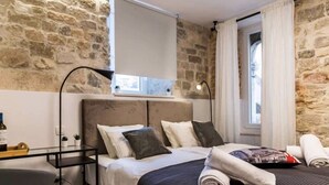 A bedroom with a stone wall, a bed with pillows and a blanket, a bedside table with a bottle, and a window with curtains. - 7heaven Luxury Rooms Palace (Split)