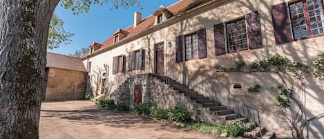 Un edificio de piedra tradicional con una escalera que lleva a una puerta de madera, rodeado por un patio de grava y un árbol grande.