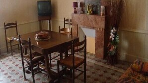 Een eetruimte met een houten tafel en stoelen, een televisie op een meubelstuk, en een decoratieve schoorsteenmantel met een beeldje en bloemen.