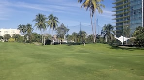A golf course with a well-maintained green, palm trees, and a modern building in the background. - Ocean front Resort  StudioMaster  (2A&2K) with Golf (2x1) in Mayan Palace in AP (Acapulco)