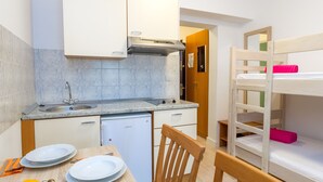 Cocina compacta con lavamanos, estufa y refrigerador pequeño. Se observa una mesa de comedor con platos y una silla. En el fondo, se ve una litera con ropa de cama rosa.