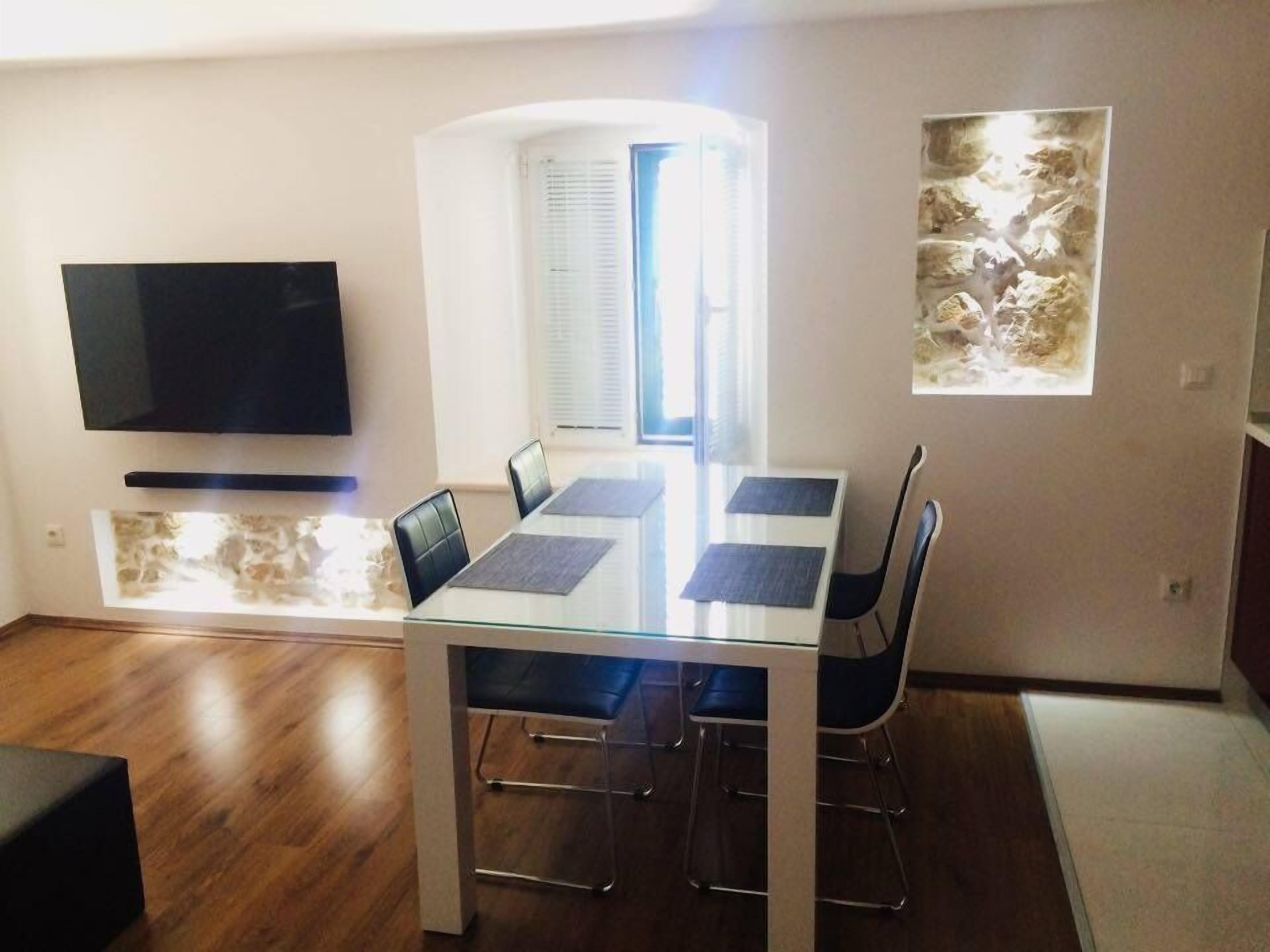 A modern dining area with a glass table, metal chairs, and a flat-screen TV mounted on the wall.