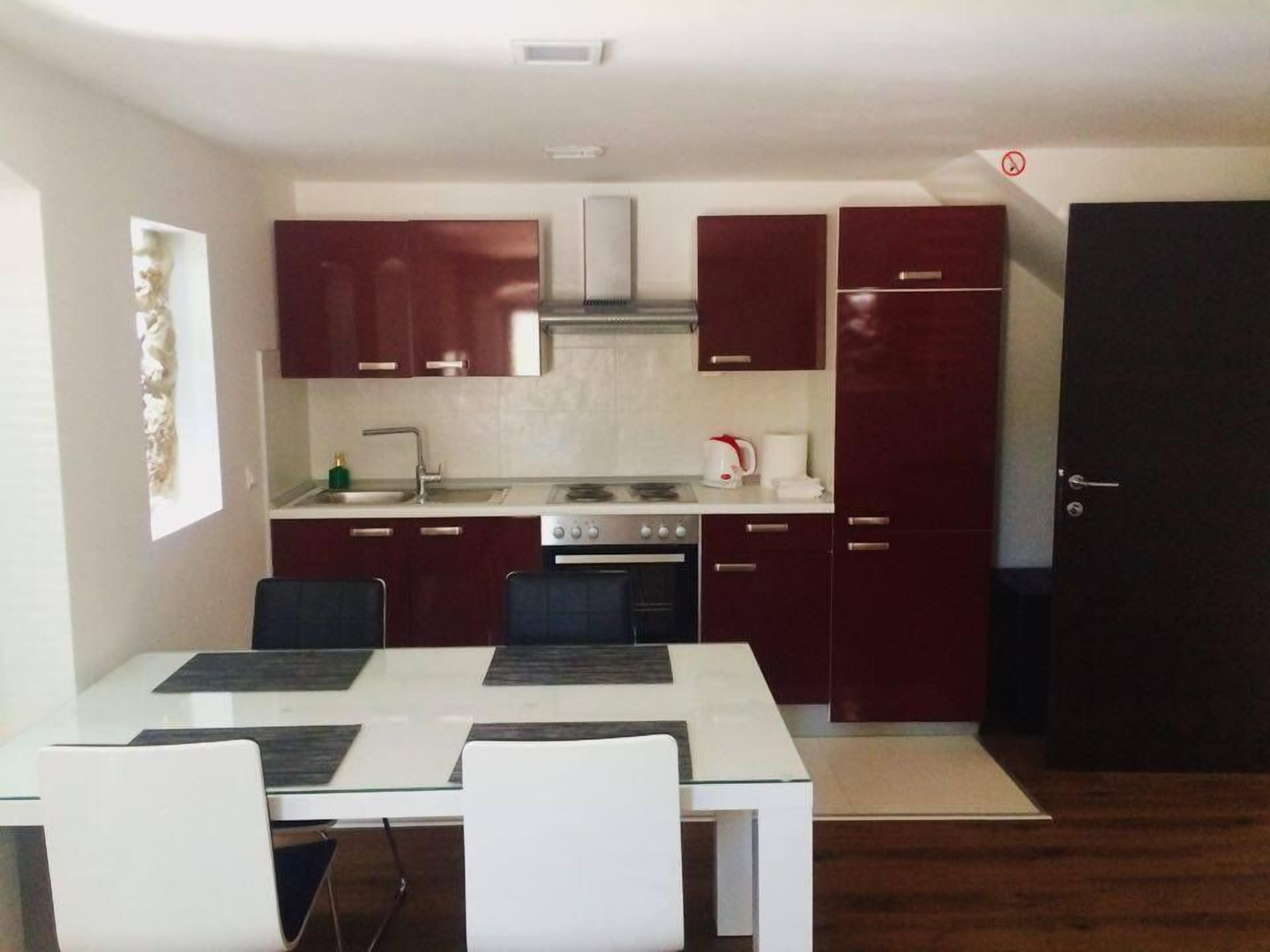 A compact kitchen with dark red cabinets, a white countertop, a stove, a sink, and a black refrigerator. There is a dining table with two chairs.