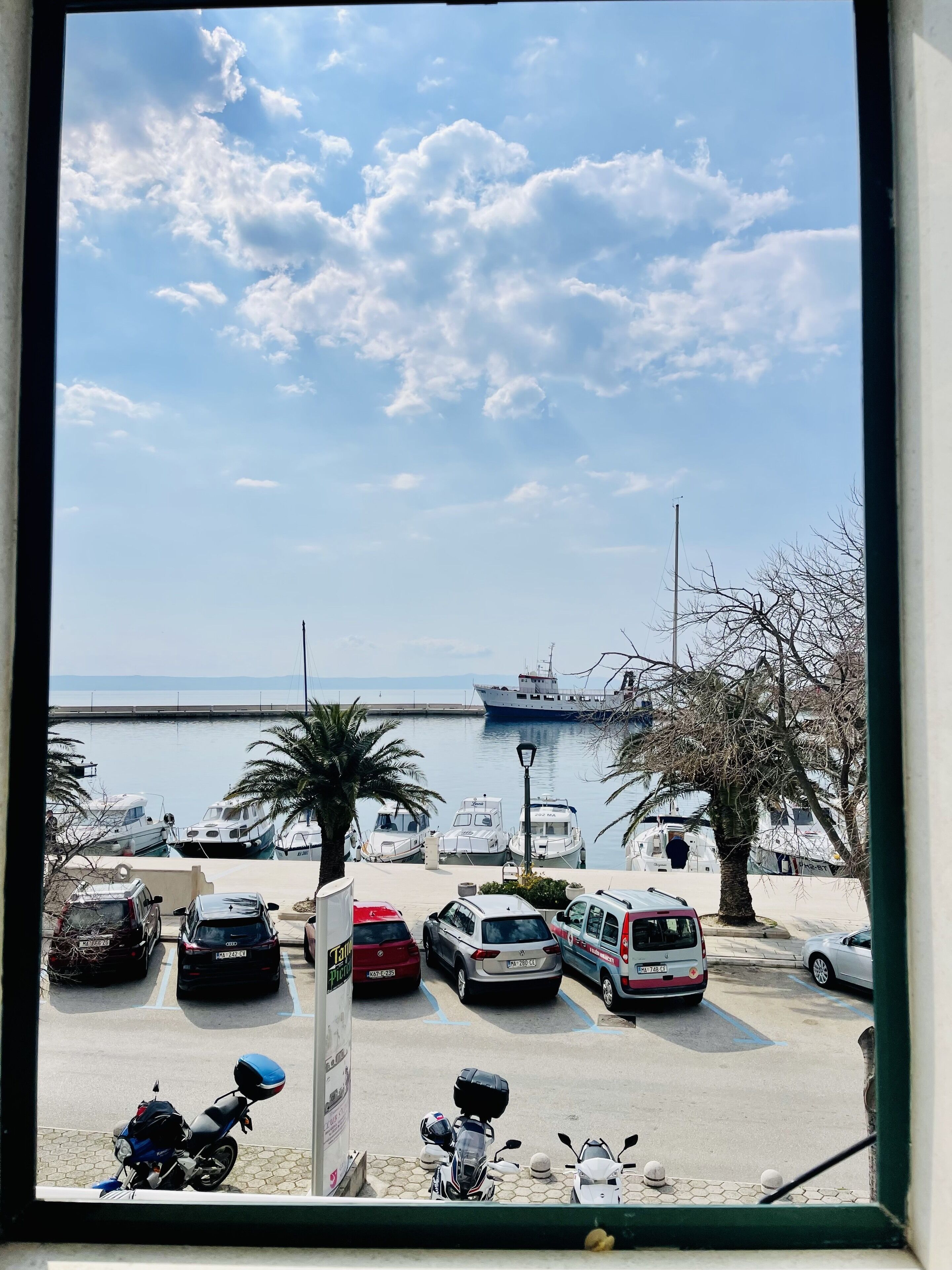 Un puerto deportivo con barcos amarrados, un puente al fondo y un aparcamiento con coches y motocicletas.