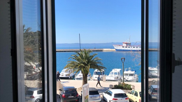 Une vue depuis une fenêtre donnant sur une marina où des bateaux sont amarrés, un piéton qui passe et des voitures garées.