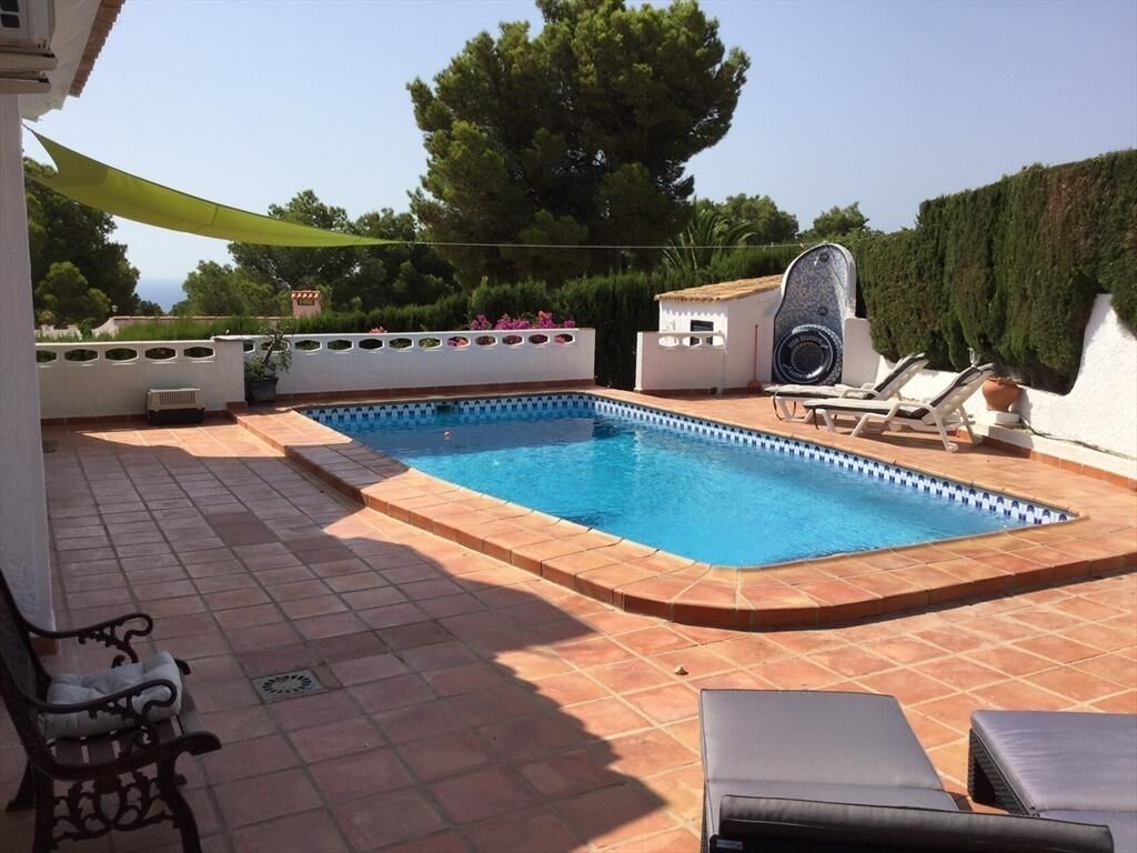 Ein rechteckiger Pool mit Fliesenverkleidung, umgeben von Liegestühlen und einem kleinen Gebäude mit dekorativer Fassade.