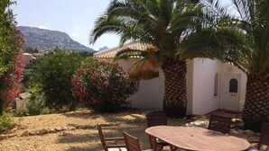 A patio with a round table and chairs, surrounded by palm trees and flowering plants, with a mountain in the background. - Santa Belgica, Altea (Altea)