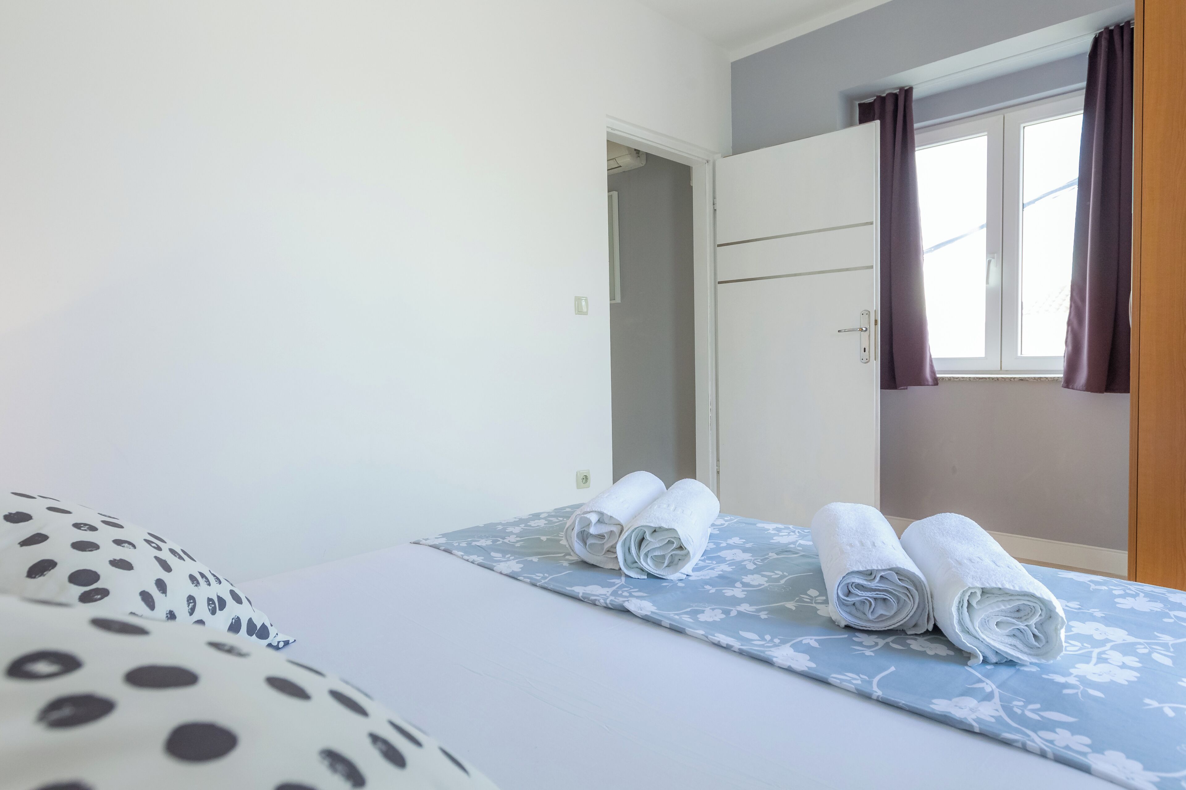 Een slaapkamer met een bed, twee opgerolde handdoeken, een raam met gordijnen en een deur.
