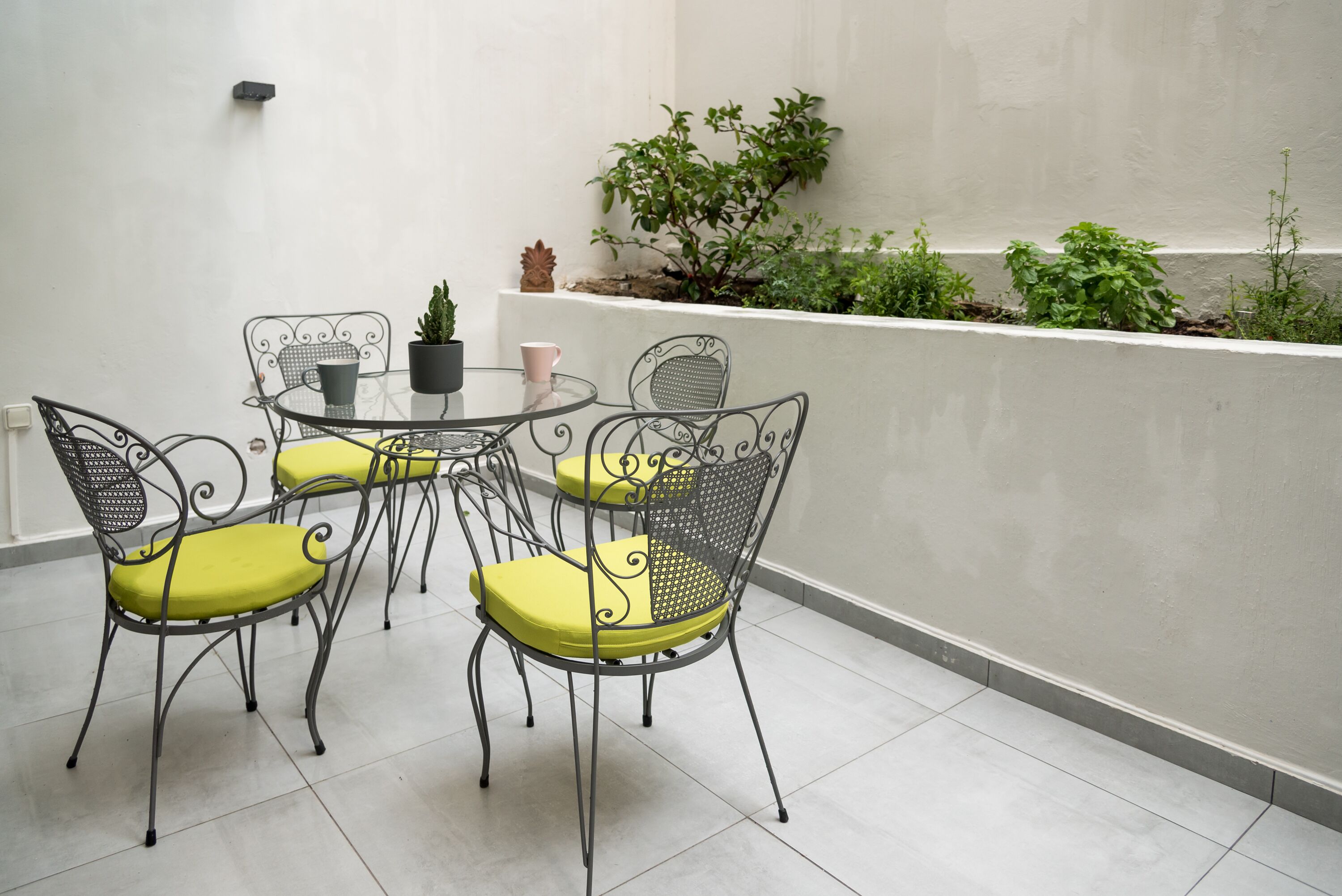 Une petite terrasse extérieure avec une table ronde et quatre chaises, entourée d’un mur blanc et d’un jardinier planté de verdure.