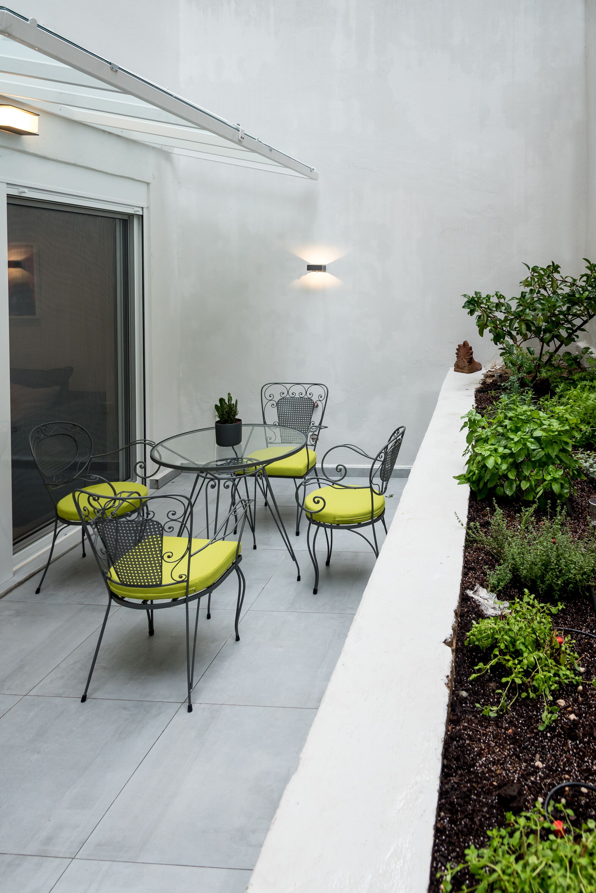 A patio with a round table and four chairs, a potted plant, and a small garden bed with various plants.