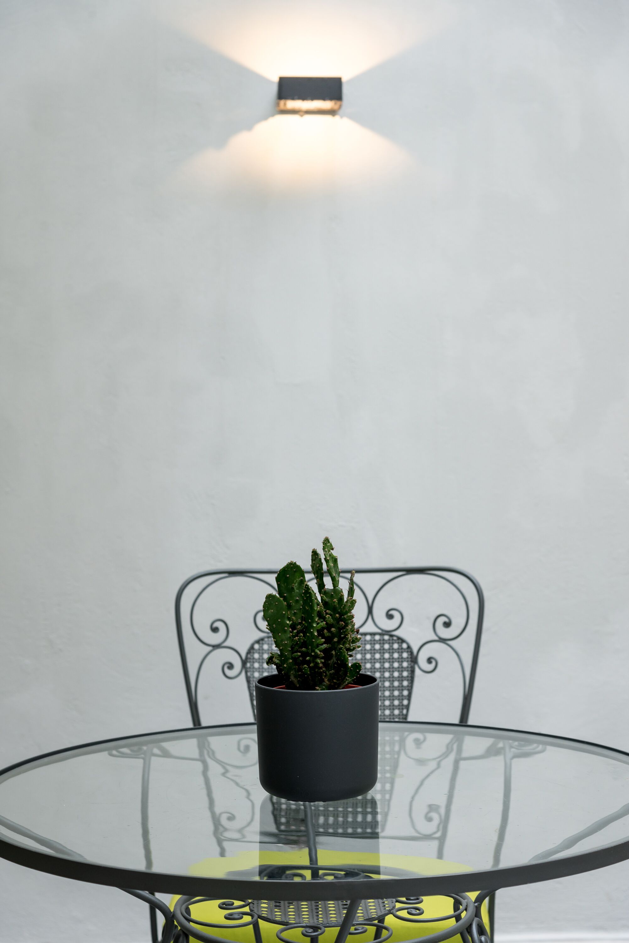Ein runder Glastisch mit einem Blumentopf, ein Metallstuhl mit verziertem Rücken und eine Wandleuchte.