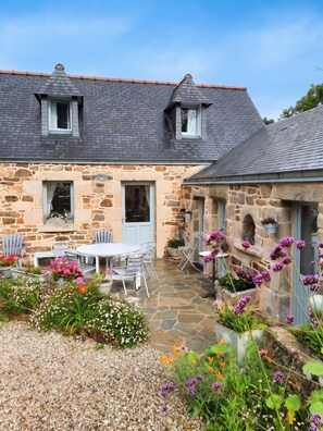 A stone house with a tiled roof, a patio with outdoor furniture, and a garden with various plants and flowers. - Homerez - House in Cléden-Cap-Sizun (Cléden-Cap-Sizun)
