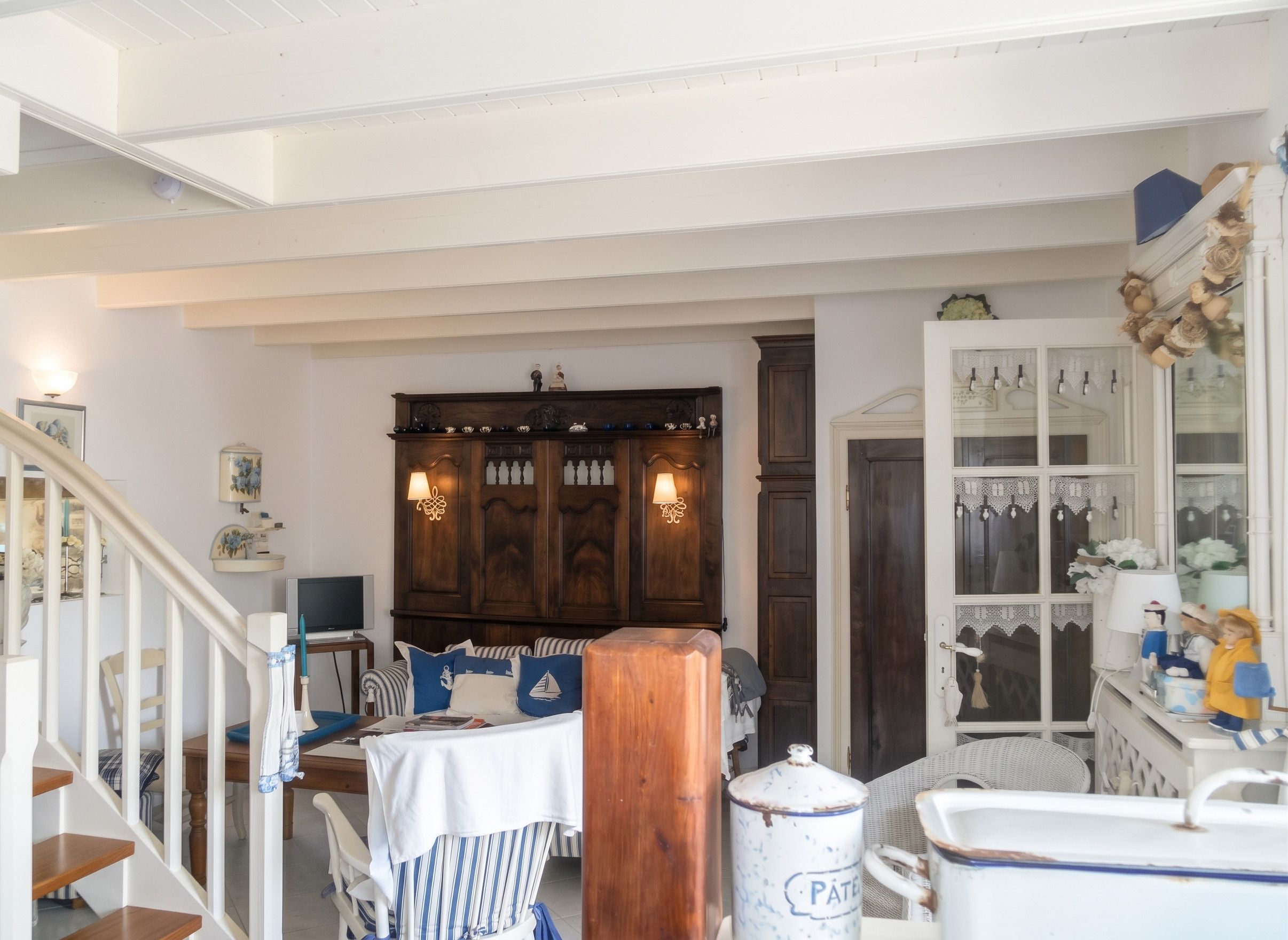 A room with a staircase, a wooden cabinet, a table with striped tablecloth, and a white cabinet with glass doors.