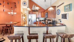 A kitchen with a central island, wooden bar stools, hanging decorative items, and a clock on the wall. - Look Out: Soundfront Condo, Pool, Great Views! (Nags Head)