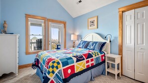 A bedroom with a large bed, two windows, a white dresser, a bedside table, and a door. - Look Out: Soundfront Condo, Pool, Great Views! (Nags Head)