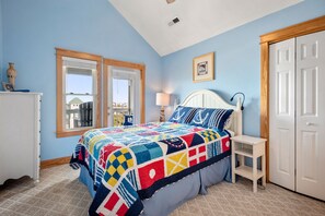 A bedroom with a large bed, two windows, a white dresser, a bedside table, and a door.