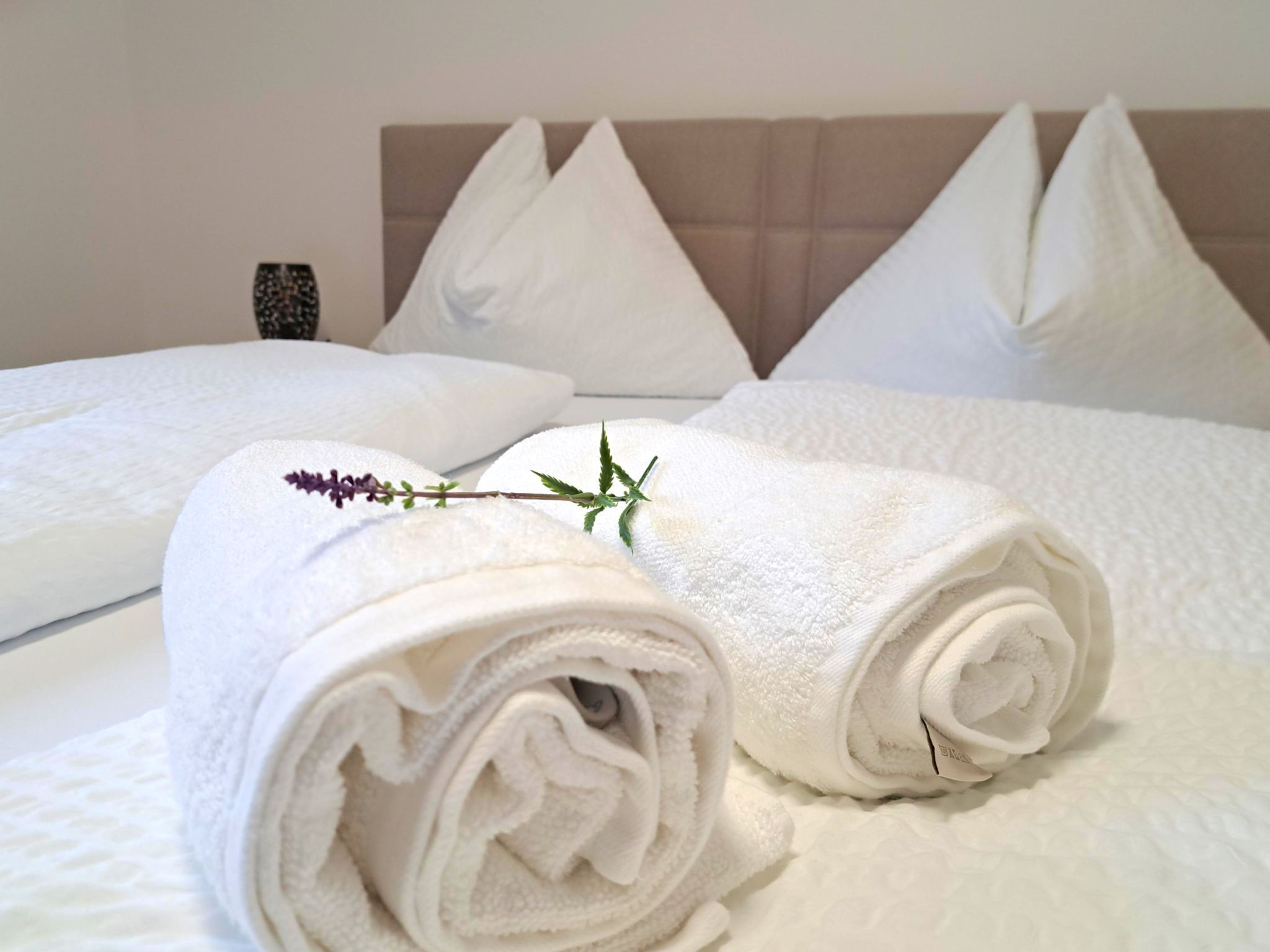A neatly made bed with white linens and rolled towels, a headboard with triangular pillows, and a small decorative item on the nightstand.