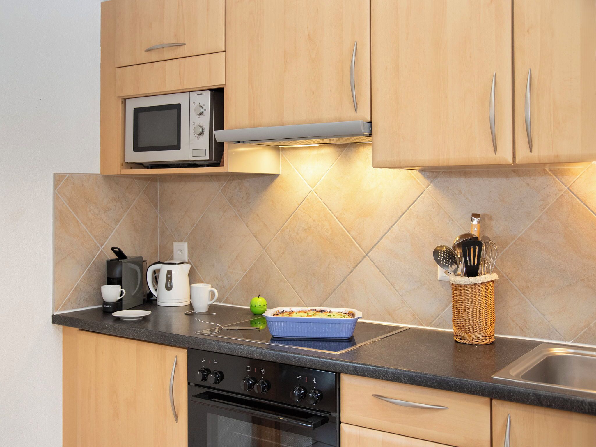 A kitchen with wooden cabinets, a microwave, a kettle, a coffee maker, and a dish in a blue baking dish.