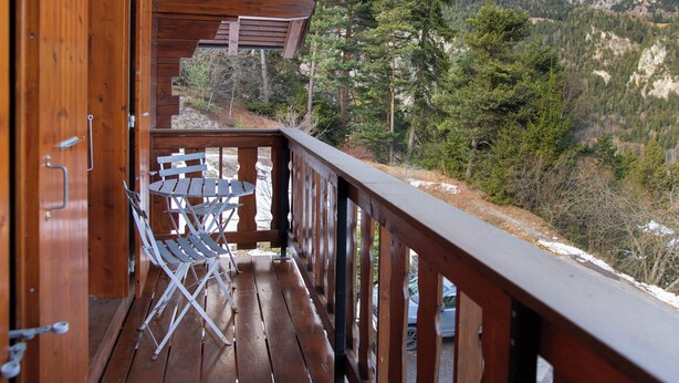 Una terraza de madera con una mesa y sillas, con vista a un paisaje montañoso con árboles y un pico nevado.
