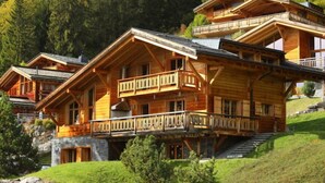 Un chalet en bois à plusieurs étages, doté de balcons, situé sur une colline, avec un mur de pierres et de la verdure.
