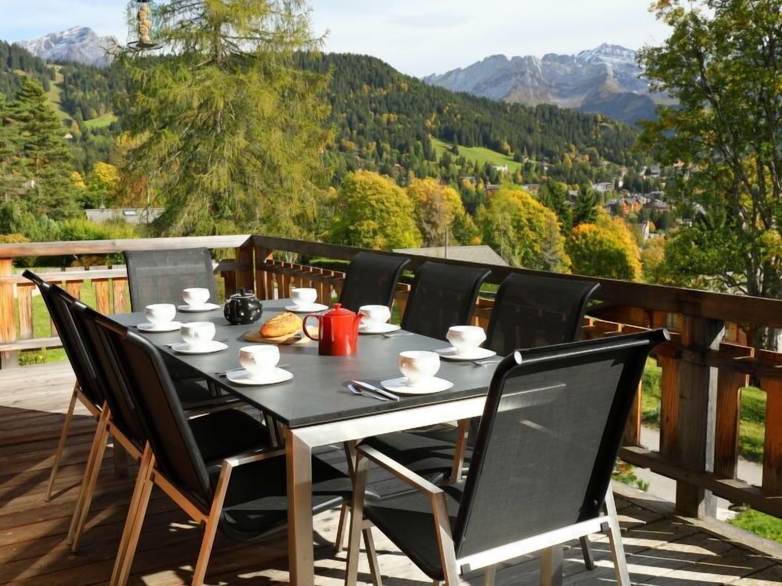 Ein Esstisch ist gedeckt mit weißen Tellern, Tassen und einer roten Teekanne. Eine Terrasse bietet Blick auf Berge und Bäume.