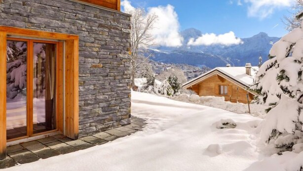 Une cabane en bois aux murs de pierre, un paysage enneigé et des montagnes en arrière-plan.