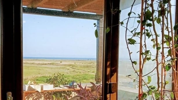 A room with a view of the sea, a wooden ceiling, and a window with a view of the sea and greenery outside.