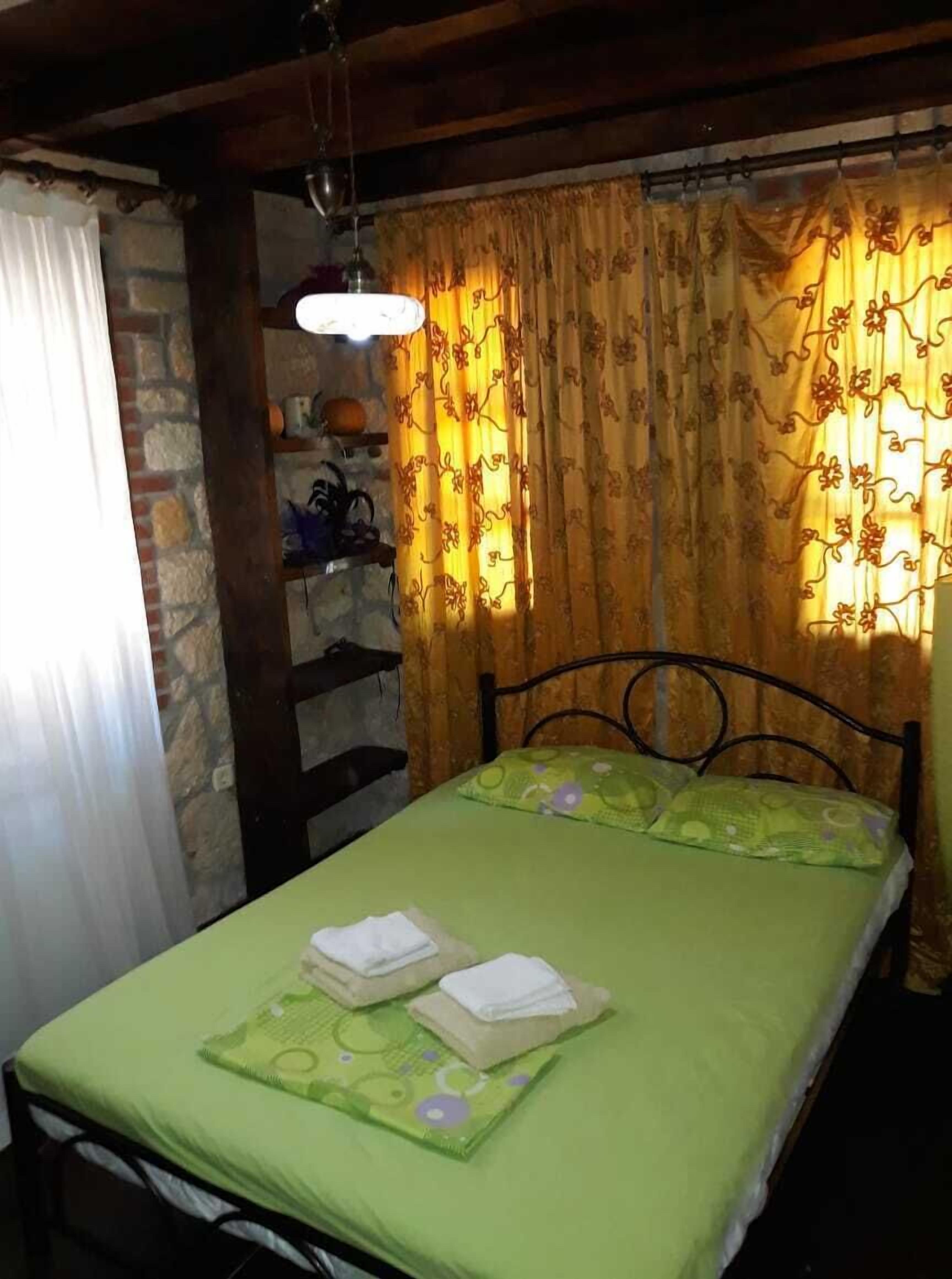 Una cama individual con colcha y almohadas verdes, cabecera de madera, una pequeña estantería con objetos decorativos y una ventana con cortinas estampadas.